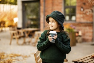 Cute little girl drinking hot cocoa from paper cup in outdoor cafe or backyard. Autumn concept