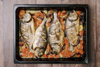 Baked sea bream with vegetables on a baking sheet stands on a wooden table. Top view.
