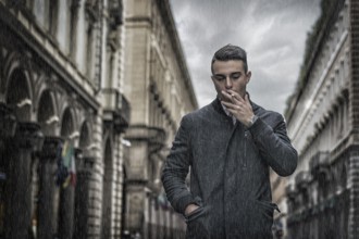 Full length of young man in coat walking under rain along the city street holding cigarette