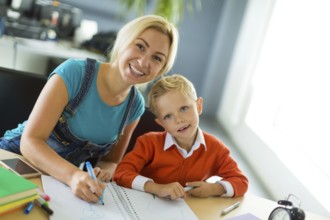 Young blonde caucasian kid in orange shirt and his mother sit at the table with colorful folders