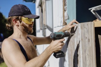 A man in overalls and sunglasses using a power drill on wooden siding outdoors