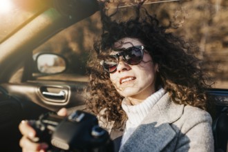 A relaxed woman with sunglasses holding a camera in a sunny convertible ride