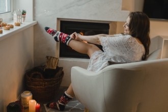 The young girl is sitting in a cozy room by the fireplace. The girl is sitting in an easy chair and