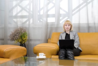 Young blonde woman sits on leather sofa with laptop, in front of glass table