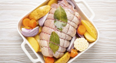 Raw wrapped rolled sliced pork in white casserole dish with potatoes, vegetables and herbs on