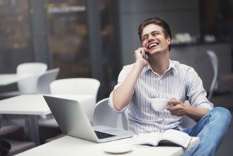 Handsome caucasian young man with phone working on laptop and smiling while enjoying coffee in cafe