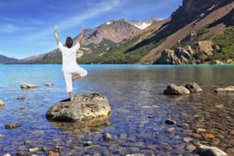 Yoga on the lake. Mature woman in white performs asana tree, standing on a rock