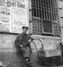 France in the 1950s. A man in the street is sitting having a cigarette. He leans on a primitive