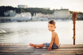 The theme is a child and summer beach holiday. A small Caucasian boy sits sideways on a wooden pier