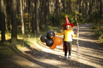A girl in a witch costume and hat on a broom with orange and black balloons is playing in the