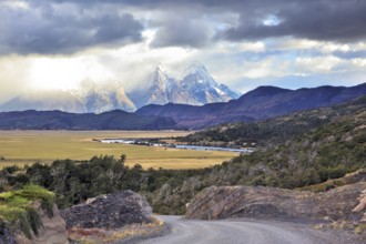 The road between turned yellow hills and river goes to snow-covered black rocks. Patagonia.