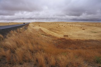 Big highway among autumn fields after harvesting. The cloudy sky and the big stacks of wheat