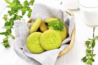 Green mint cookies on napkin in wicker basket, milk in a glass and a jug on white wooden board