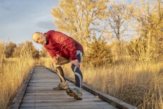 Senior male runner is stretching on a boardwalk trail, fall sunset at Riverbend Ponds, popular