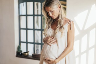 Schöne schwangere Frau, die ihren Bauch berührt und am Fenster steht