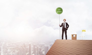 Young caucasian businessman standing on house roof and holding go green sign. Mixed media