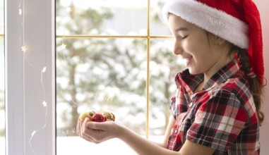 Girl in a Santa hat sits on the windowsill of a house near the Christmas tree and holding glass