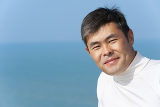 Handsome asian young man looks into camera smiling, blue sea and sky