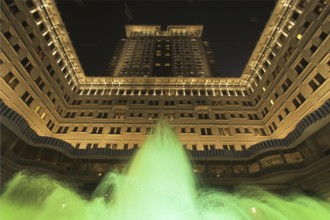 The jets of a fountain illuminated by green light before an enormous architectural complex in Hong