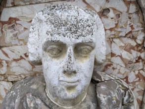 Weathered stone bust with classical features and detailed carving against a textured background,
