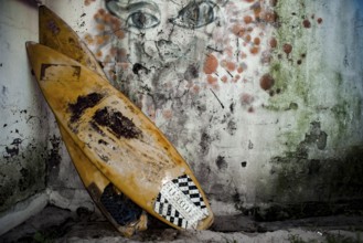Two old surfboards lie stacked against a mouldy wall with faded, bad, graffiti on the wall