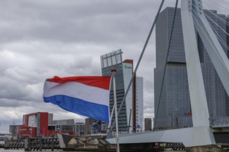 Dutch flag waving in front of a bridge and modern buildings under a cloudy sky, Rotterdam,