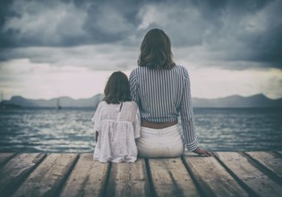 Woman and little girl sitting on the wooden pier near the sea