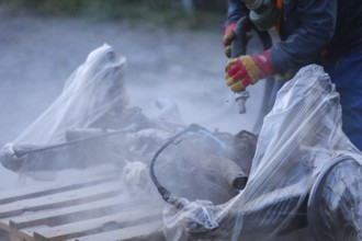 A worker in protective gloves sanding car parts covered in plastic, producing dust