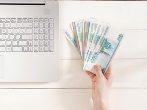 Hand holding russian rubles near laptop on white wooden background. Top view or flat lay. Finance