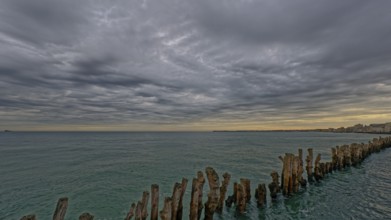 Calm sea with a row of wooden posts extending into the water under a dramatic cloudy sky,