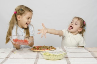 Two little girls at a table prepared pizza. One with a whimsical view stretches a hand to a plate