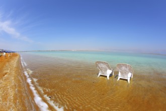 Two white beach armchairs stood side by side in the water at the beach. Dead Sea, Israel