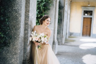 Smiling bride in a dress with a bouquet of pink flowers sat down near a pillar in a vaulted room.