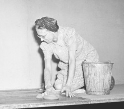 Elderly cleaning lady is on her knees scrubbing floorboards dressed in a classic cleaning coat and