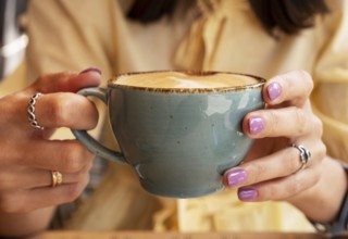 Woman in yellow clothes holding blue cup of cappuccino with both hands, close-up
