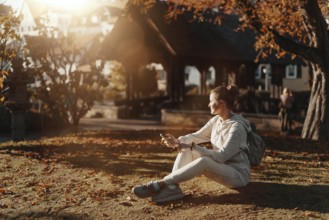 Young fashionable teenage girl with smartphone in park in autumn sitting at smiling. Trendy young