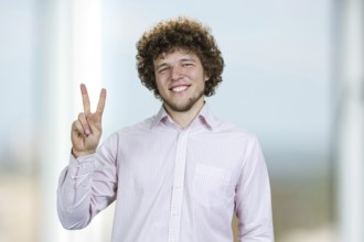 Portrait of a happy young man with curly hair shows victory gesture sign. Indoor window in the