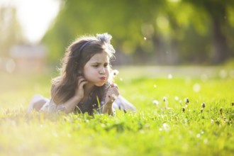 The little girl lays on a grass and blowing dandelion on the summer day