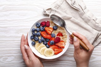 Top view of female hands holding bowl with oatmeal porridge with fruit and berries and spoon on