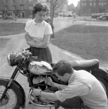 In the 1950s. A man on his Puch motorcycle with a woman looking at him while he checks the fuel