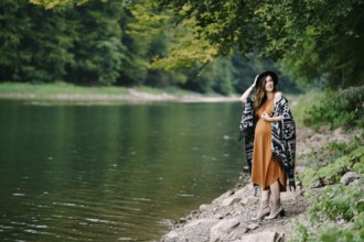 Pregnant woman stands on a rocky lake shore in the forest.