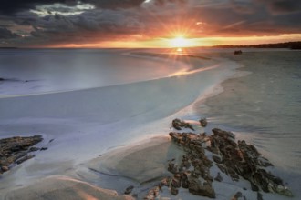 Picturesque La Pelosa beach in Sardinia at sunrise with a sunburst and rocks in the foreground