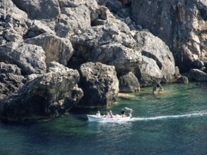A small boat sails along the rocky coast in clear blue water, rocky blue sea with green bushes and