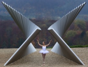 Ballerina in white ballet costume in elegant pose in front of modern sculpture in autumnal