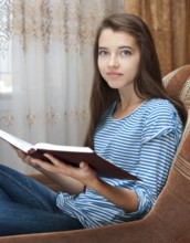 A young caucasian teenage girl reads a book sitting in an armchair