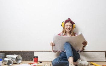 Happy girl in protective headphones sitting on floor with paper blueprint. Home remodeling after