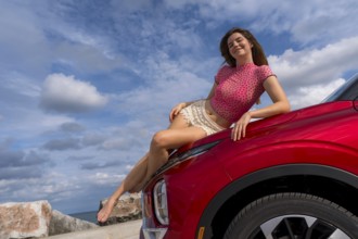 A gorgeous redhead model sits confidently on the hood of her car, soaking in the summer breeze as