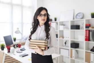 Beautiful young girl stands in the office and holds a stack of books in her hands