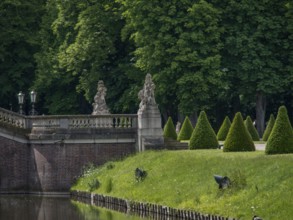 A stone bridge with statues leads over a river, flanked by neat hedges and lush trees, nordkirchen,