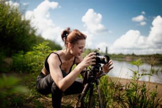 A woman smiles as she adjusts her camera by the lakeside under a blue sky
